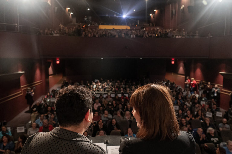 Two people on stage speaking to a full audience