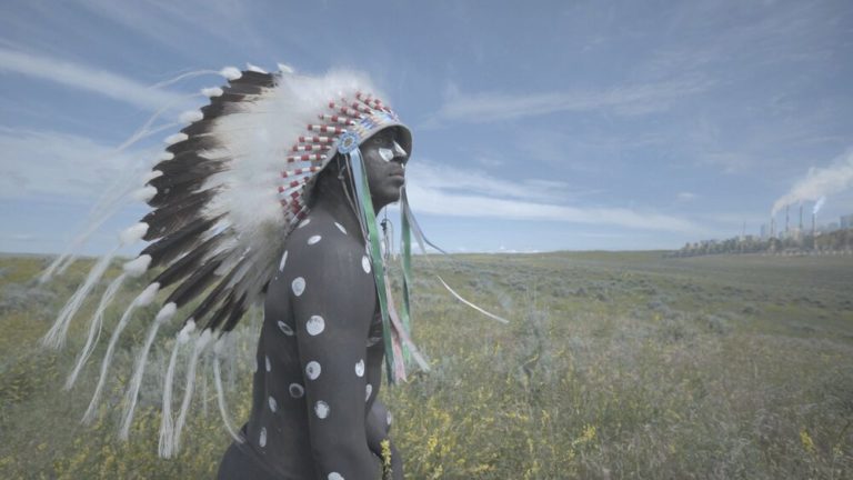 A video still of an indigenous person in body paint and a headdress.